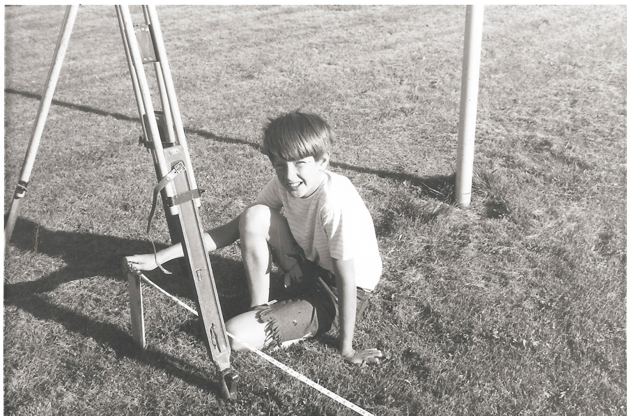 A young person sitting on grass next to a tripod. They are wearing a striped shirt and shorts. There is a string on the ground in front of them.