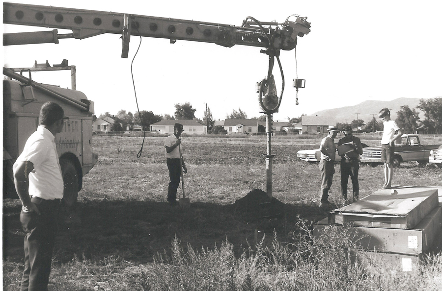 A group of five men stand in an open field next to a large piece of machinery. One man stands on a platform, while another holds a clipboard and talks to two men nearby. A fifth man stands a short distance away, examining the scene. In the background, there are houses, cars, and distant mountains. The side of a vehicle has the partial text "Electrical" visible.