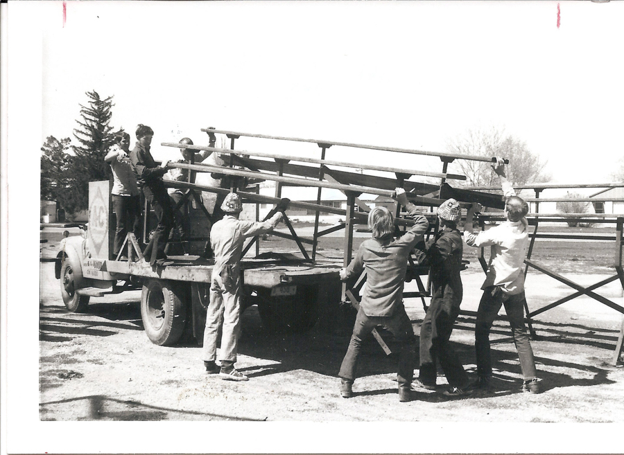 A group of seven people, some wearing hats and work clothes, are working together to lift a large, metal framework onto a flatbed truck. The truck has a logo with the letters "H-C" and a phone number for Northrup on its side. There are trees and buildings visible in the background.