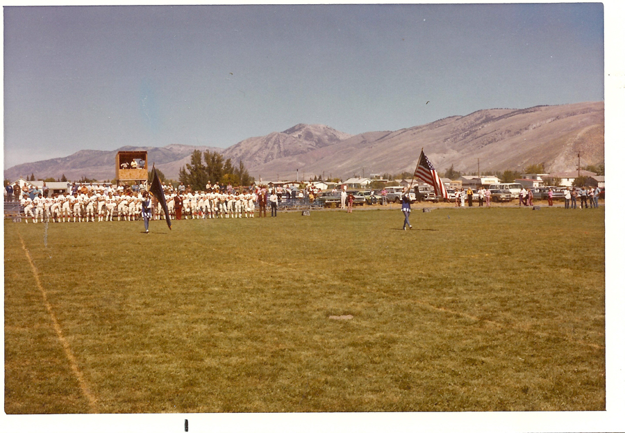 A group of people in sports uniforms stand on a grassy field with mountains in the background. Two individuals carry large flags, including an American flag, towards the center. Spectators are seated on bleachers, with more people standing near vehicles in the distance. A wooden platform or booth is visible with people observing the scene. The field is marked with numbers, such as "40" and "50."