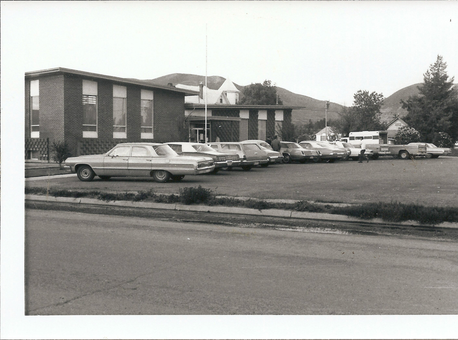 A row of vintage cars parked in front of a rectangular brick building with large windows. In the background, there are hills and several trees. The scene includes a person walking near the cars and a white bus parked further back.