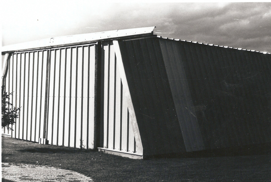 A large, angled metal structure with vertical panels stands on a grassy area. The structure appears to be divided into sections, with one section slightly darker than the other. The sky above is cloudy.