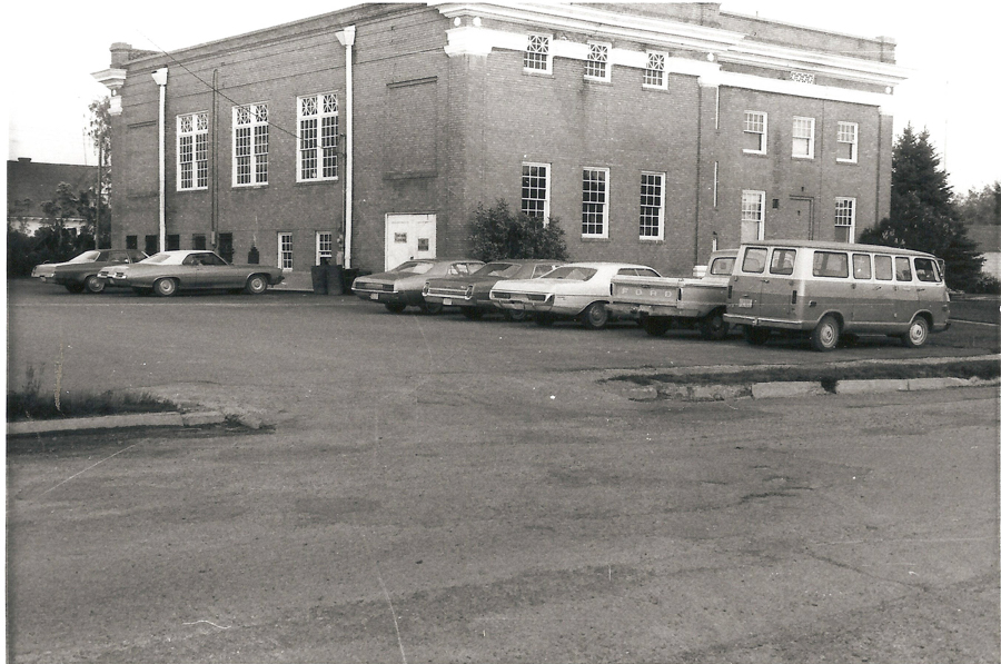 A parking lot with several cars and a van parked in front of a large brick building. The building has tall windows and a decorative roofline. The side of a Ford pickup truck can be seen, and there are some trees and shrubbery in the background.