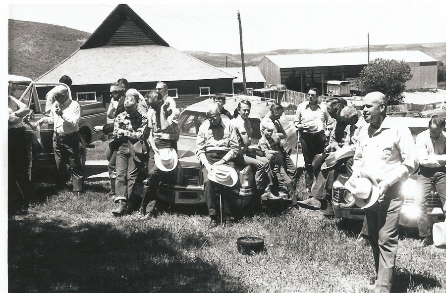 A group of men stands and sits on cars in a grassy area near a barn. They are wearing casual clothing, and some hold hats. A boy is among them, sitting on a car. There are other buildings and vehicles in the background.