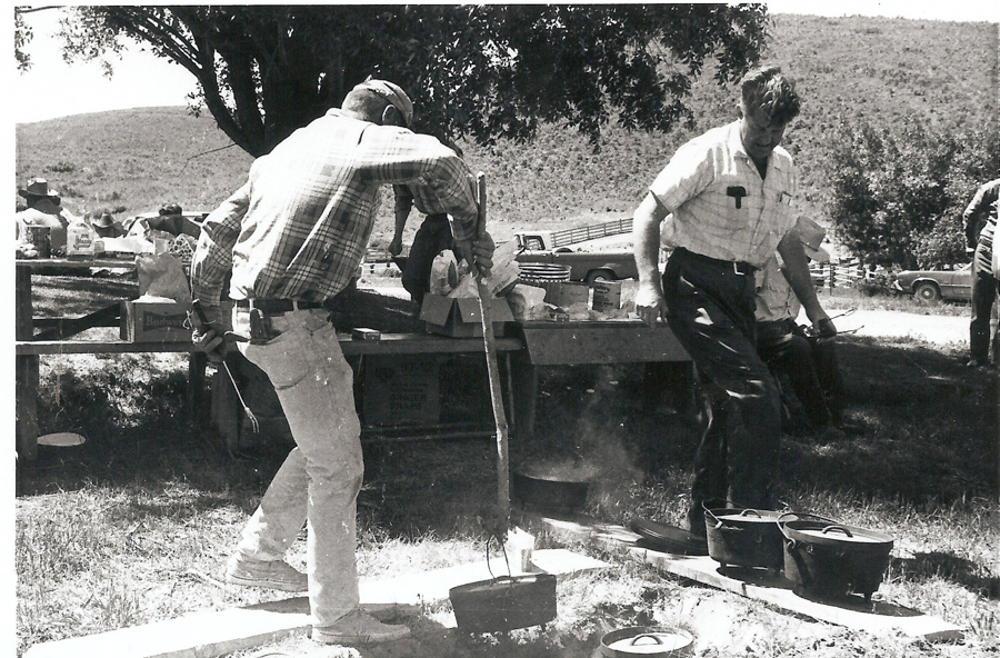 A man wearing a cap and plaid shirt is tending to a pot over a fire using a long stick, while another man in a short-sleeved shirt stands nearby, holding utensils. There are several pots and a wooden table nearby with supplies on it, including visible boxes with text that reads "Budweiser" and "Stoker's Sliced Beets." In the background, people are seated at a picnic table under a tree, and a car is parked further behind them.