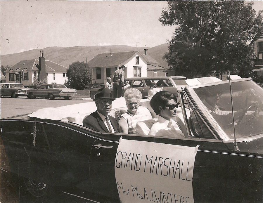 A man with a hat, an elderly woman, and a young woman wearing sunglasses are seated in a convertible. The convertible has a sign on the side that reads "GRAND MARSHALL Mr. & Mrs. A.J. Winters." In the background, there are several houses, parked cars, and a tree.