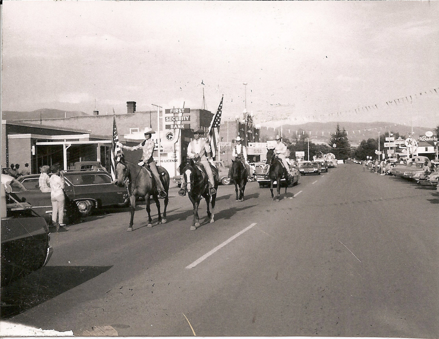 People riding horses on a street during a parade, carrying American flags. Cars are parked along the road, and spectators stand nearby. A sign for "First Security Bank" is visible, along with other businesses. Mountains can be seen in the distance.
