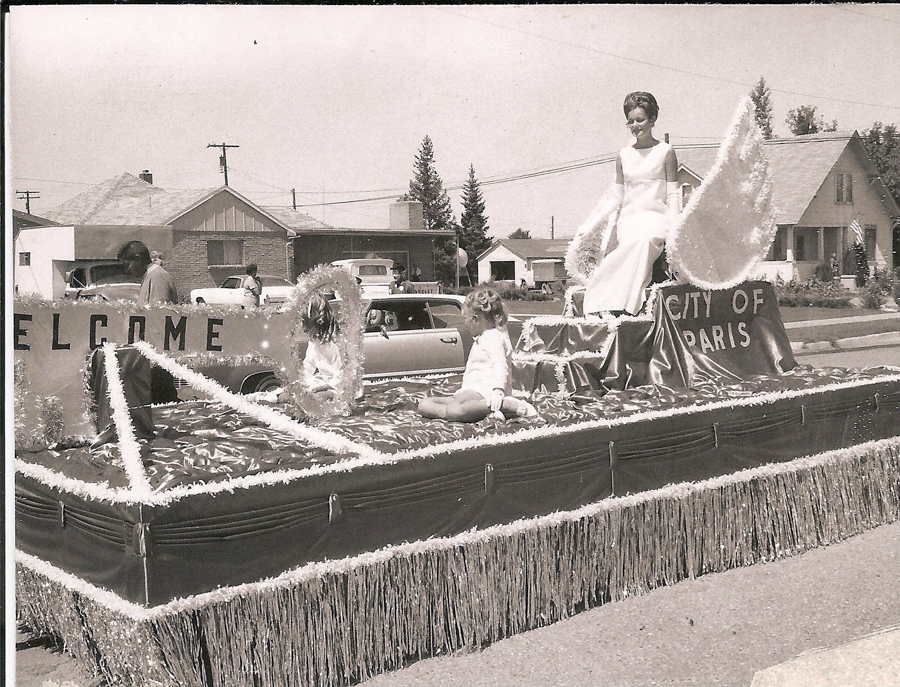 A woman in a long dress sits on a float decorated with tinsel and fabric. The float has large letters that say "WELCOME" and "CITY OF PARIS." Two children are sitting on the float, surrounded by tinsel. Houses and cars are in the background.
