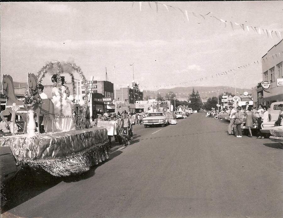 A parade scene on a street with a float featuring a person in a gown and two horse figures. People in costumes walk beside the float holding a banner. Shops line the street with visible signs including "First Security Bank" and "Western Auto." Cars are parked along the sides, and spectators are gathered watching the parade. Bunting is strung across the street.