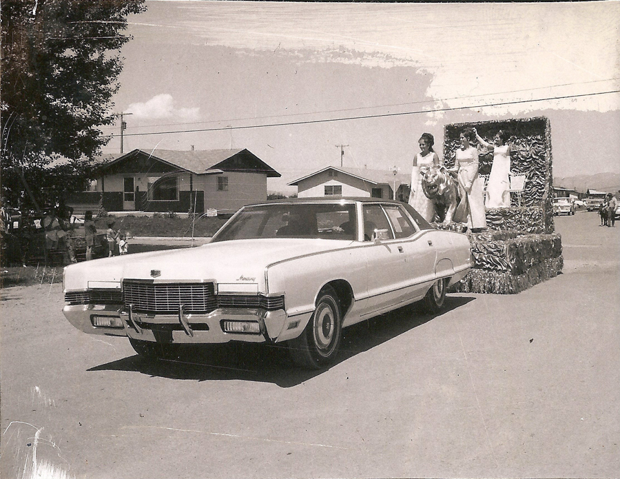 A car is driving in a parade, towing a decorated float. Three women in long dresses stand on the float, which features what appears to be a large model tiger. Houses and trees are visible in the background, and a small group of people is gathered along the roadside.