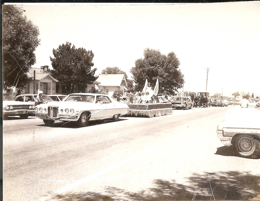 A street scene featuring a parade with several cars and a float. The float is decorated and carries people, some of whom are holding flags. Trees and houses line the street. In the background, there are more vehicles and onlookers. The setting suggests a community event or celebration.