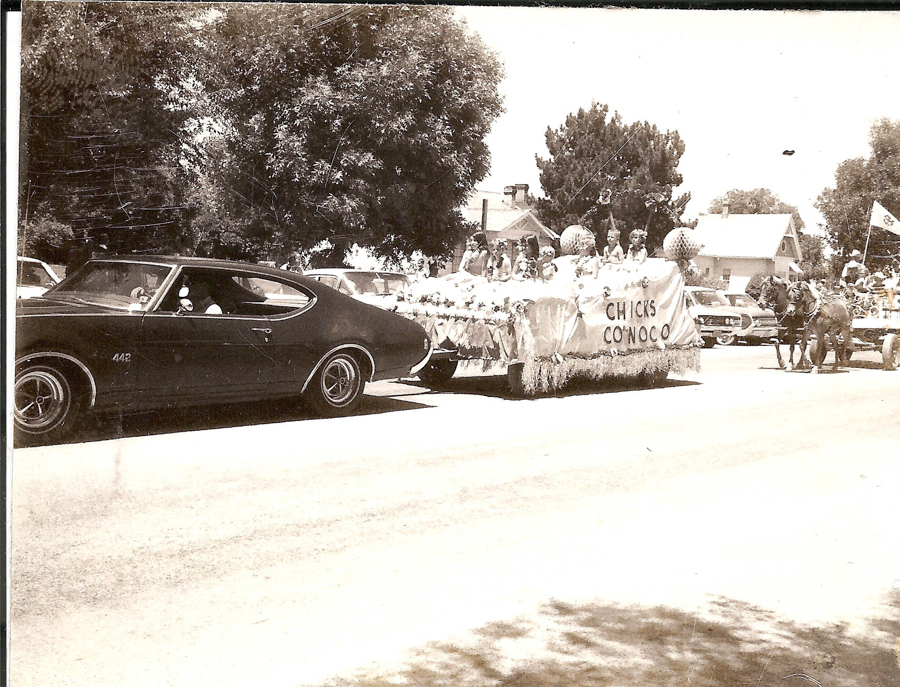 A parade with a classic car in the foreground, followed by a float carrying girls in dresses beneath a sign that reads "CHICK'S CONOCO." In the background, a horse-drawn carriage carries additional participants, with houses and trees lining the street.