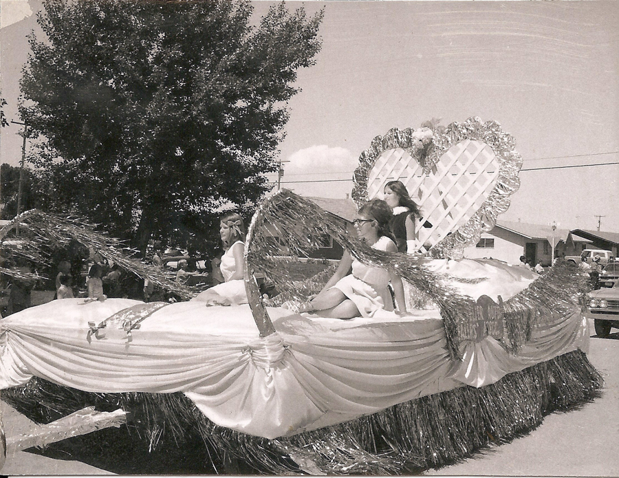 A parade float decorated with draped fabric, tinsel, and a large heart backdrop. Two women in formal dresses are seated on the float, one wearing glasses. In the background, people stand under trees and near buildings, watching the parade. A partial view of a car can be seen on the right side.
