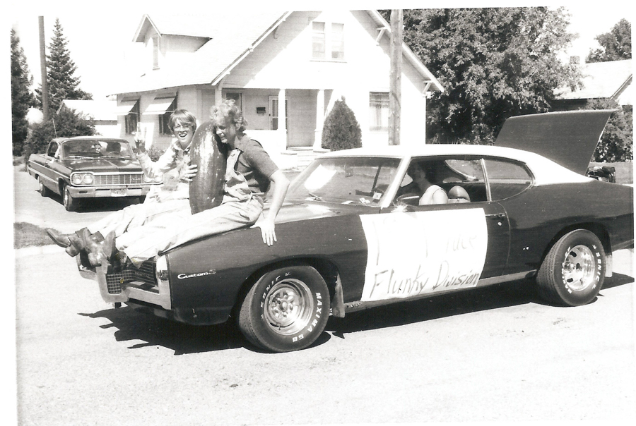 Two people sitting on the hood of a car, holding a large object between them. The car has a sign on its side that reads "1st Place Funky Division." Another person is sitting inside the car. In the background, there is a house and another parked car on the street.