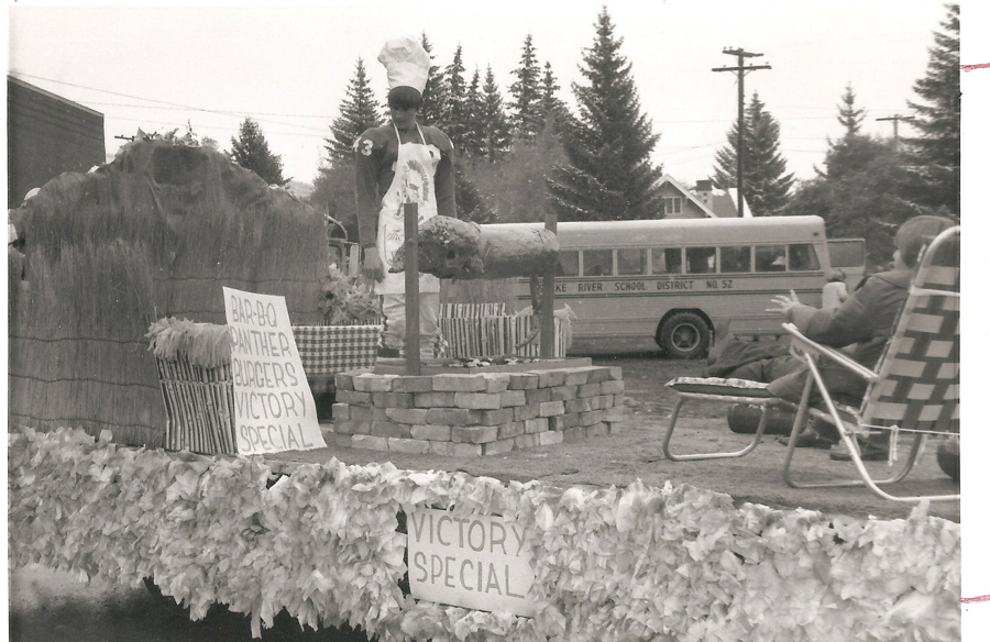 A person wearing a chef's hat and apron stands on a float. The float displays a model animal on a spit over a mock brick platform. Signs on the float read "BAR-B-Q PANTHER BURGERS VICTORY SPECIAL" and "VICTORY SPECIAL." In the background, there's a school bus labeled "KOKE RIVER SCHOOL DISTRICT NO. 57" and trees. A person in a folding chair is seated nearby.