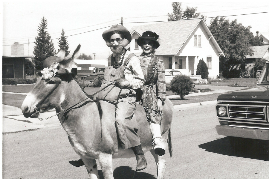 Two people riding a donkey on a residential street. The person in front is wearing a hat, glasses, and overalls, while the person behind is dressed in a patterned dress and hat. A car is partially visible on the right, and houses and trees line the street in the background.
