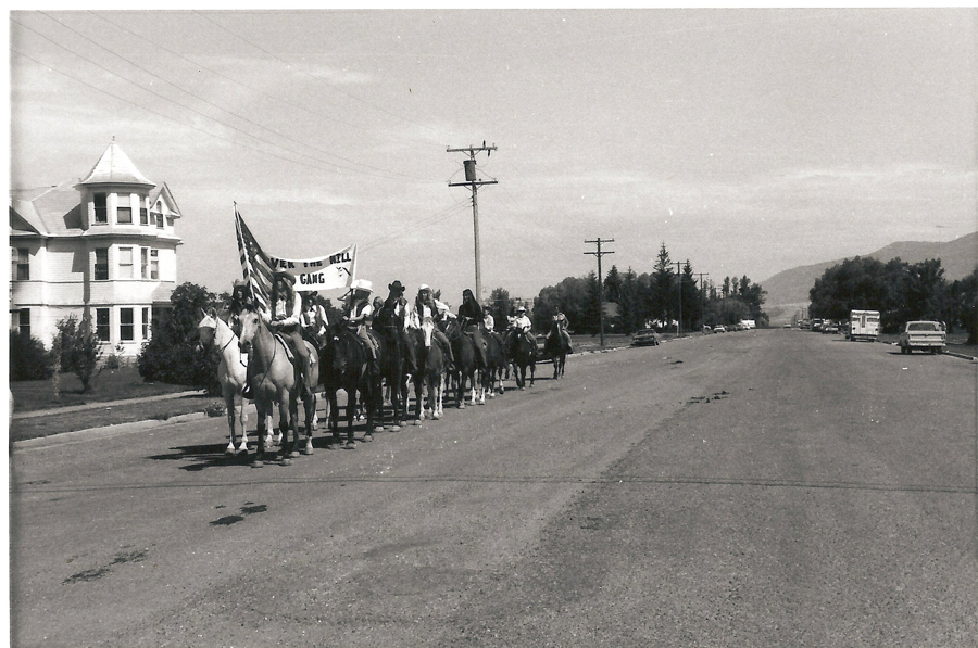 A group of people on horseback rides down a street. They carry a banner that reads "Over the Hill Gang." An American flag is also visible. The street has a large house on one side and a few cars parked along it. Trees and power lines are in the background.