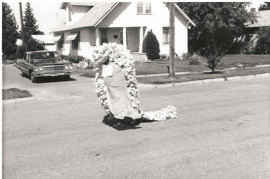 A person in a costume that looks like a large caterpillar or dragon, made of fabric and ruffles, is moving down a residential street. There is a vintage car parked on the side of the street, and a house with a porch and trees in the background.