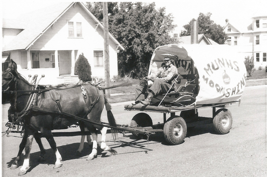 A man wearing a hat sits on a horse-drawn cart resembling a small covered wagon, labeled "Munns Moonshine." The cart is pulled by two horses. Houses and trees are visible in the background along a street.