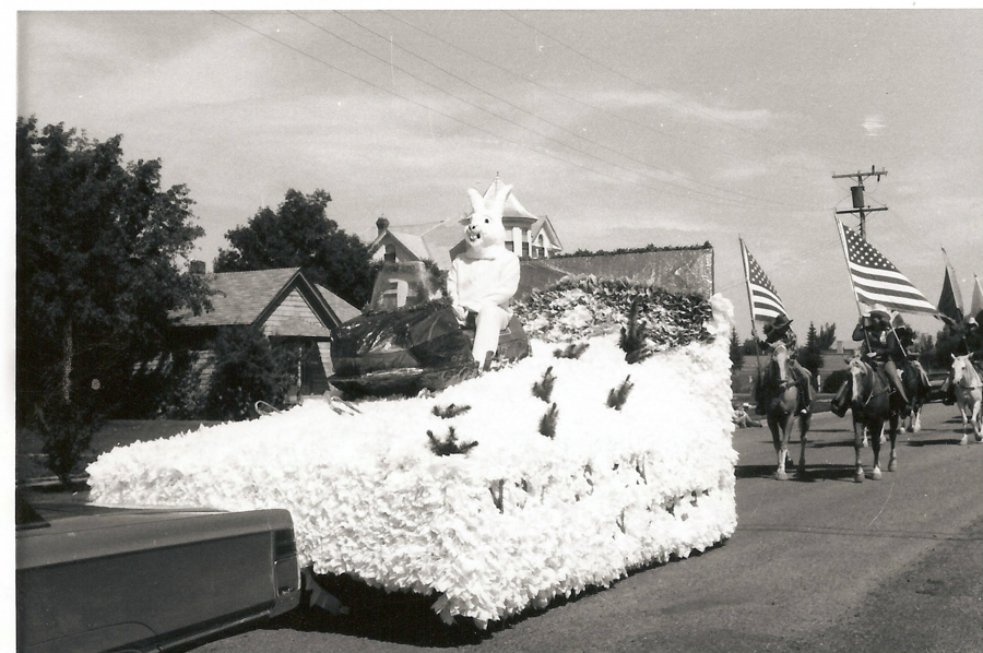 A person dressed in a rabbit costume sits on a float covered in decorative materials. The float is part of a parade with people on horses carrying American flags. Houses and trees are visible in the background.