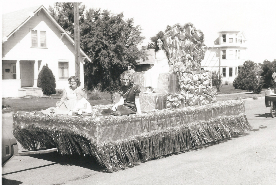 Three women sitting on a decorated parade float with fringes and ornamental designs. The word "WESTERN" is partially visible on the side of the float. They are in a residential area with houses and trees in the background.