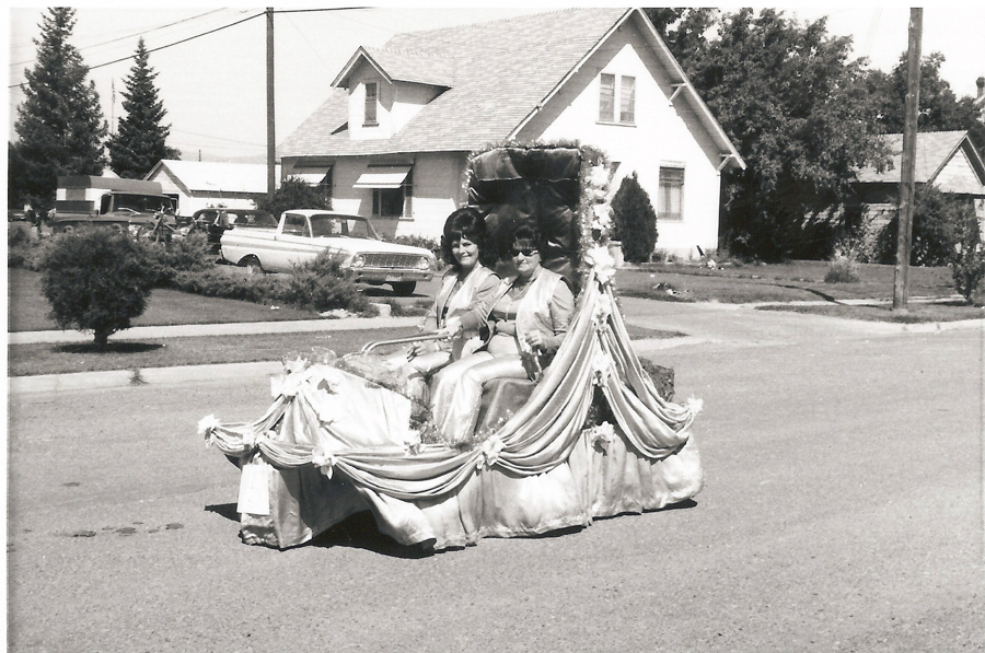 Two women sitting on a float decorated with draped fabric and flowers, riding on a residential street. Houses, a parked car, and trees are in the background.