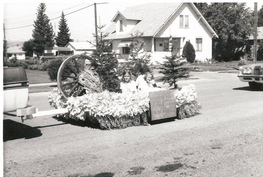 Two children are sitting on a decorated float being towed by a vehicle in a street. The float is adorned with flowers, small evergreen trees, and a large wagon wheel. A sign on the float reads "BUCKAROO BREAKFAST OUR SUNDAY STAMPEDE FLOAT BOYS AND GIRLS." Houses, cars, and utility poles are visible in the background.
