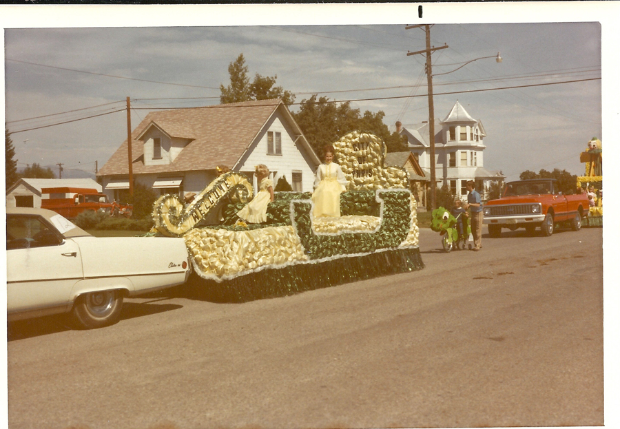 A street scene featuring a parade with a decorative float. The float, adorned in green and beige materials, carries two women wearing yellow dresses. The word "Welcome" is displayed prominently on the float. A white car is seen in the foreground, and a red truck follows behind. A man stands beside a child on a decorated bicycle. Residential houses with trees and a large utility pole are visible in the background.