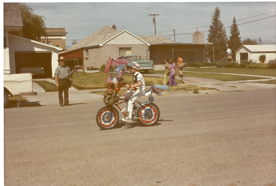 A child wearing a helmet rides a bicycle decorated with balloons and patriotic colors on a residential street. An older man stands nearby with a hat, holding an object. In the background, three people are walking on the sidewalk near some houses and trees.