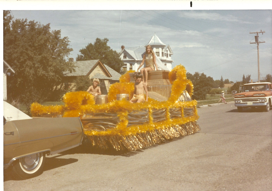 A decorated parade float on a street, featuring three children. One child, a girl, is sitting on the highest point of the float. Two boys are positioned below her on either side. The float is adorned with yellow and gold tinsel. Homes are visible in the background, along with a large house featuring a spire. A beige car and another vehicle are on the street nearby.