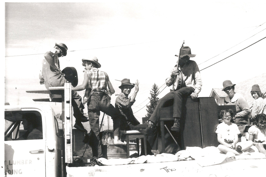 Several men wearing hats and casual clothing are on the back of a truck, with one playing a banjo and another sitting with a hat covering his face. There is a piano and a person sitting at it. Two young children sit in front, smiling and looking at the camera. The words "LUMBER" and "ING" are partially visible on the truck door.
