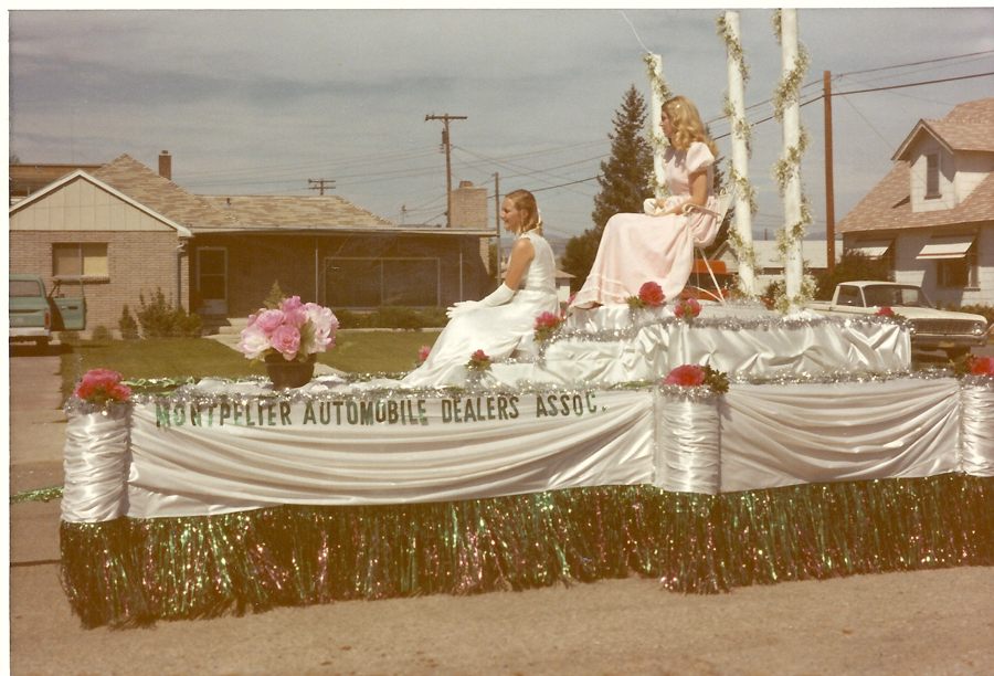 Two women in formal dresses sitting on a decorated parade float. The float is adorned with flowers and has signage reading "MONTPELIER AUTOMOBILE DEALERS ASSOC." The scene is set in a residential neighborhood with houses, a car, and a van visible in the background.