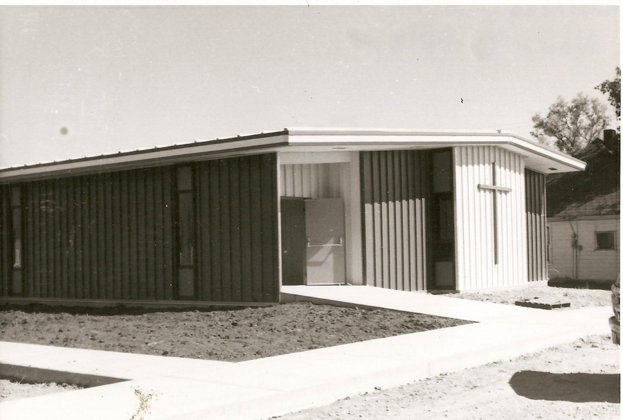 A single-story building with a modern design featuring vertical paneling. A large cross is mounted on one of the exterior walls. A pathway leads to the entrance of the building. A small house is visible in the background on the right side.