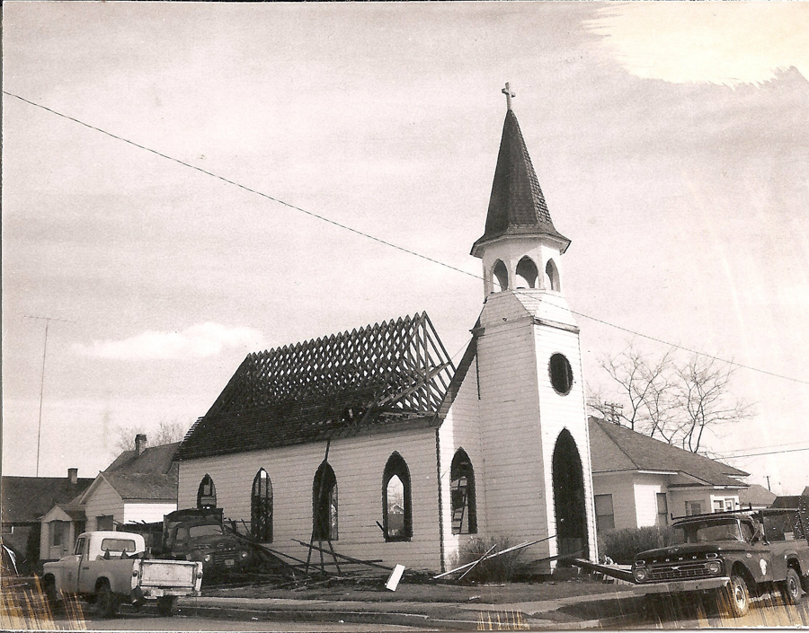 A church building with roof under construction, featuring a tall steeple with a cross on top. Several trucks are parked around the church. Nearby, there are small houses and leafless trees. Broken wooden structures and debris are visible around the scene.