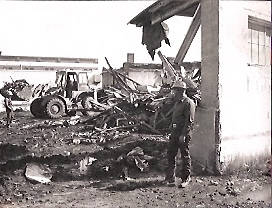 A person wearing a hard hat stands next to a partially demolished building. In the background, a large construction vehicle is visible near a pile of rubble and debris. The scene appears to be a construction or demolition site with exposed beams and materials. The ground appears uneven with dirt and debris scattered around.