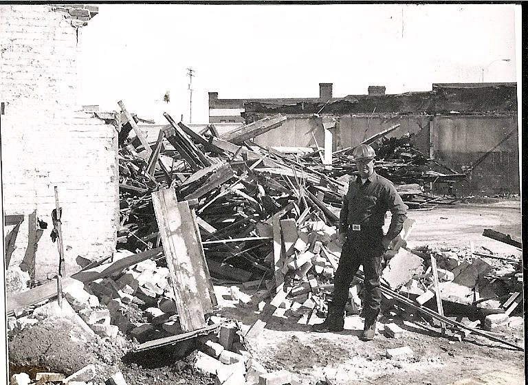 A man wearing a hard hat stands amidst rubble and debris. Piles of wooden beams and bricks are scattered around. In the background, partially standing walls of a damaged building are visible.