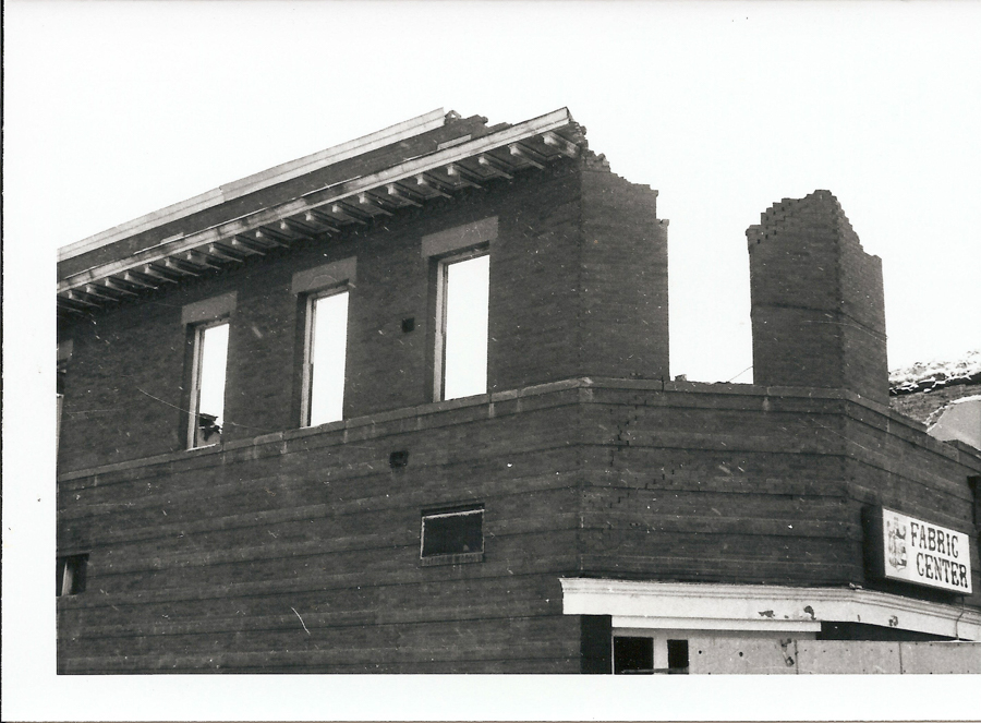 A partially demolished brick building with three tall windows. The top part of the structure shows damaged corners. A sign on the lower right side reads "Fabric Centre" with a partially visible logo.