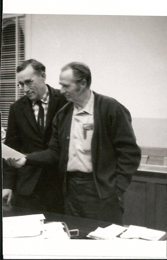 Two men stand close together, looking at a piece of paper one is holding. They are in an indoor setting with a table in front of them, covered in documents and papers. One man wears a suit and tie with glasses, and the other wears a cardigan with a shirt that has a pocket containing pens. Blinds cover the window in the background.