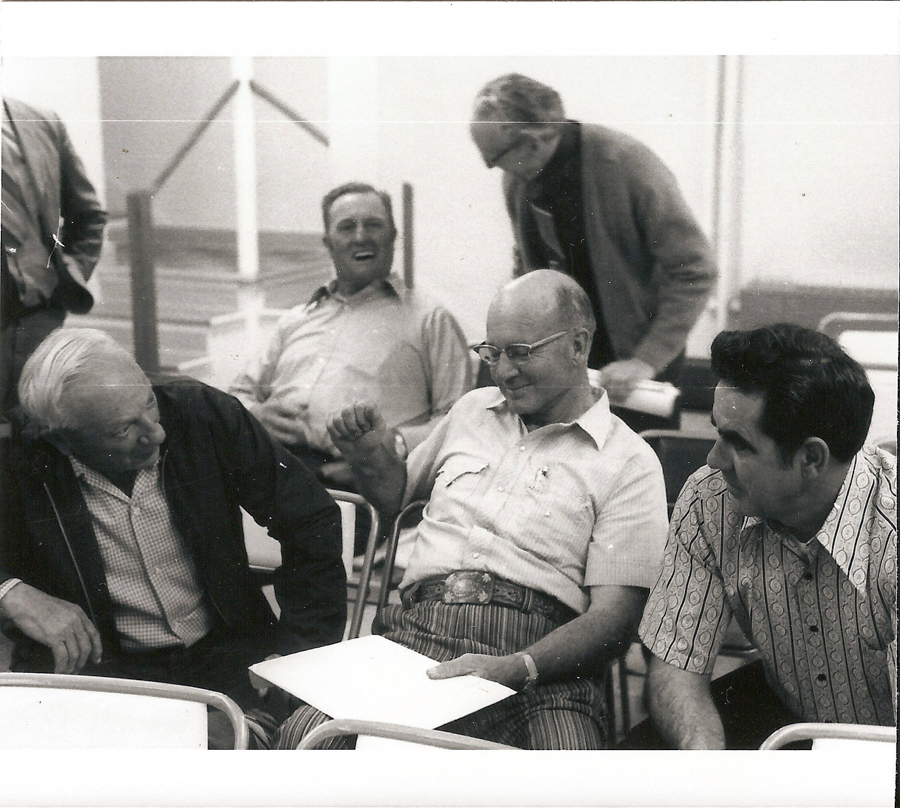 A group of five men gathered together, with three seated and two standing. The seated men appear to be engaged in conversation, one holding a sheet of paper and wearing a striped shirt. The standing men are in the background, one leaning over slightly. The setting is indoors.
