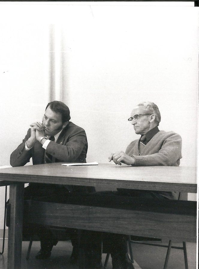 Two men seated at a table. One is resting his chin on his hand, appearing thoughtful, while the other looks ahead with arms resting on the table. The setting appears to be a simple room without decoration.