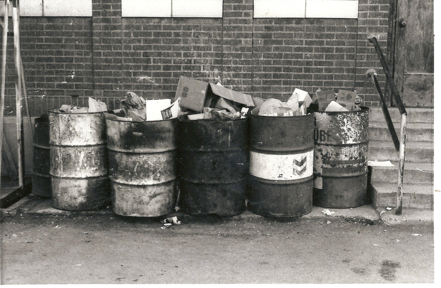 Six metal barrels filled with various pieces of cardboard and paper are lined up against a brick wall. To the right, a set of concrete steps leads up with a metal handrail. The barrels are labeled, with the word "LUBE" visible on one and a Chevron logo on another. Debris is scattered on the ground in front of the barrels.