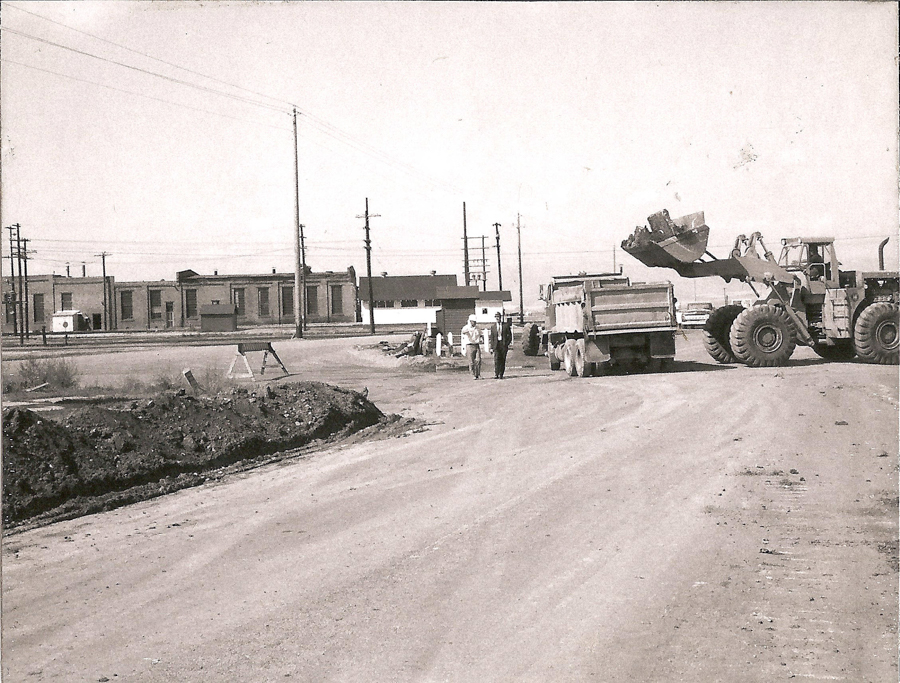 A construction scene with a large loader placing debris into a dump truck. Two workers are walking on the dirt road alongside the machinery. In the background, there are several industrial buildings and electrical poles.