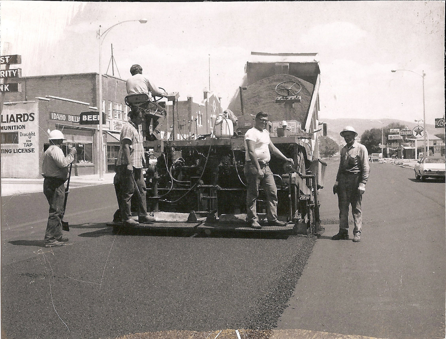 A group of workers are operating paving machinery on a street. One man is seated on the machine, while others stand nearby, some wearing hats. Buildings with signage, including "BILLIARDS" and "IDAHO BILLI" are visible in the background. A portion of a motel sign and a parked car are seen further down the street.