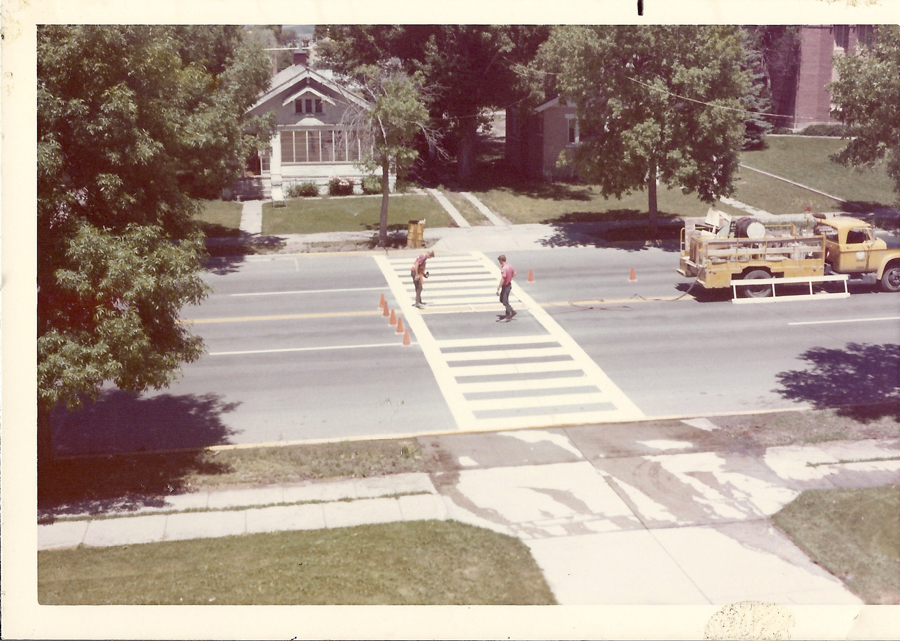 Two people are standing on a pedestrian crossing being painted on a road. Traffic cones are placed along the crossing. A yellow work truck is parked nearby. In the background, there is a house with a lawn, and trees line the street.