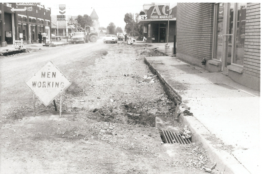 A street under construction with a dirt surface and a sign that reads "MEN WORKING." On the left side, there are buildings including a store with signage "A C FOOD MARKETS." Several vehicles and people are visible further down the street.