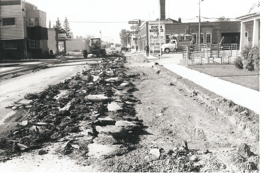 A street undergoing construction with rough gravel and debris scattered across it. On the left, there are buildings, one with a "Rich Motor Co." sign. A tractor is visible on the road, and a few cars are parked. A sidewalk and grassy area are on the right. Several signs for businesses are in the background.