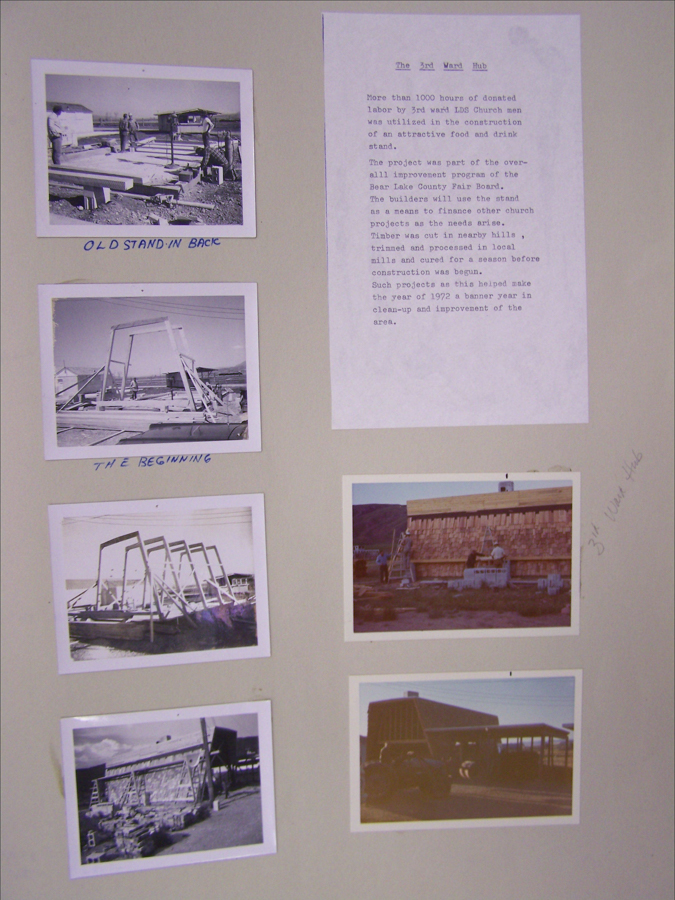 A collection of six small images is displayed on a board alongside a typed document. The images depict various stages of a construction project, with people working with timber and various structures visible. The document, titled "The 3rd Ward Hub," describes the project involving over 1000 hours of donated labor by LDS Church men for building a food and drink stand. It discusses the larger improvement program for Bear Lake County Fair Board, detailing the use of local timber and mills. Handwritten captions under the images include "OLD STAND IN BACK" and "THE BEGINNING." An additional note reads "3rd Ward Hub."
