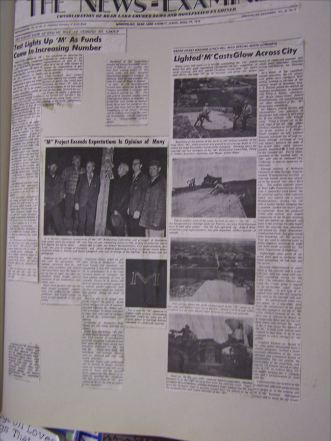 A newspaper page with multiple columns of text and several photographs. The header reads "The News Examiner" and features articles titled "Test Lights Up 'M' As Funds Come In Increasing Number" and "'M' Project Exceeds Expectations Is Opinion of Many." Another titled "Lighted 'M' Casts Glow Across City" includes three images: a group of people standing with a post, individuals working on a large letter "M" project, and the letter "M" lit up. The page also has smaller cut-out segments pasted onto it.