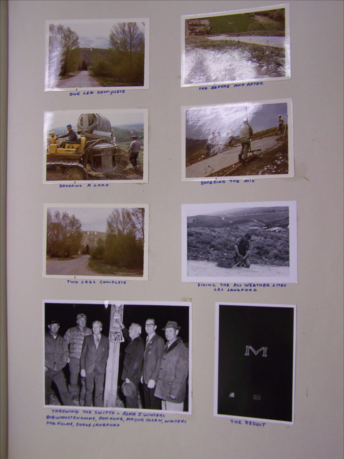 A collection of seven images with handwritten captions. 

The first image shows a dirt road lined with trees leading to a hillside captioned "One leg complete." 

The second image shows a mix being spread on the ground, captioned "The Before and After."

The third image depicts a man operating machinery next to a mixer, captioned "Dropping a load."

The fourth image shows individuals spreading a mix on a sloped path, captioned "Spreading the mix."

The fifth image features a scene similar to the first image but further along, captioned "Two legs complete."

The sixth image shows a person kneeling and working on the ground, captioned "Fixing the all weather lite, Les Lanfrod."

The seventh image features a group of men standing near a wooden post at night, captioned "Throwing the switch - Alma J. Winters; Bob Woolstenhulme, Don Hunt, Mayor Olsen, Winters, Phil Hulme, Duane Langford."

The last image is a dark background with an "M" prominently displayed, captioned "The Result."