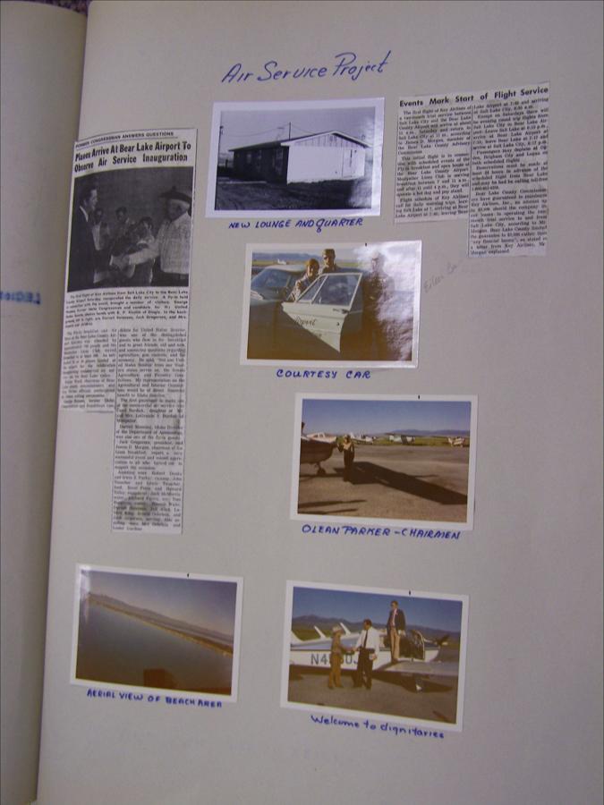 A page titled "Air Service Project" includes several photographs and newspaper clippings. 

1. A newspaper article titled "Planes Arrive At Bear Lake Airport To Observe Air Service Inauguration" with a black-and-white image of people standing together.

2. A photo labeled "New Lounge and Quarter" showing a small building.

3. Another image labeled "Courtesy Car" depicting two people standing beside a car.

4. A picture labeled "Olean Parker - Chairmen" showing a person standing near a small aircraft.

5. An image labeled "Aerial View of Beach Area" capturing a view of a coastline or beach.

6. A photo titled "Welcome to Dignitaries" with three individuals standing next to an aircraft, with one person on the wing.